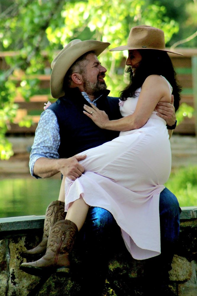A couple in hats laughing, she sits in his arms on a stone wall